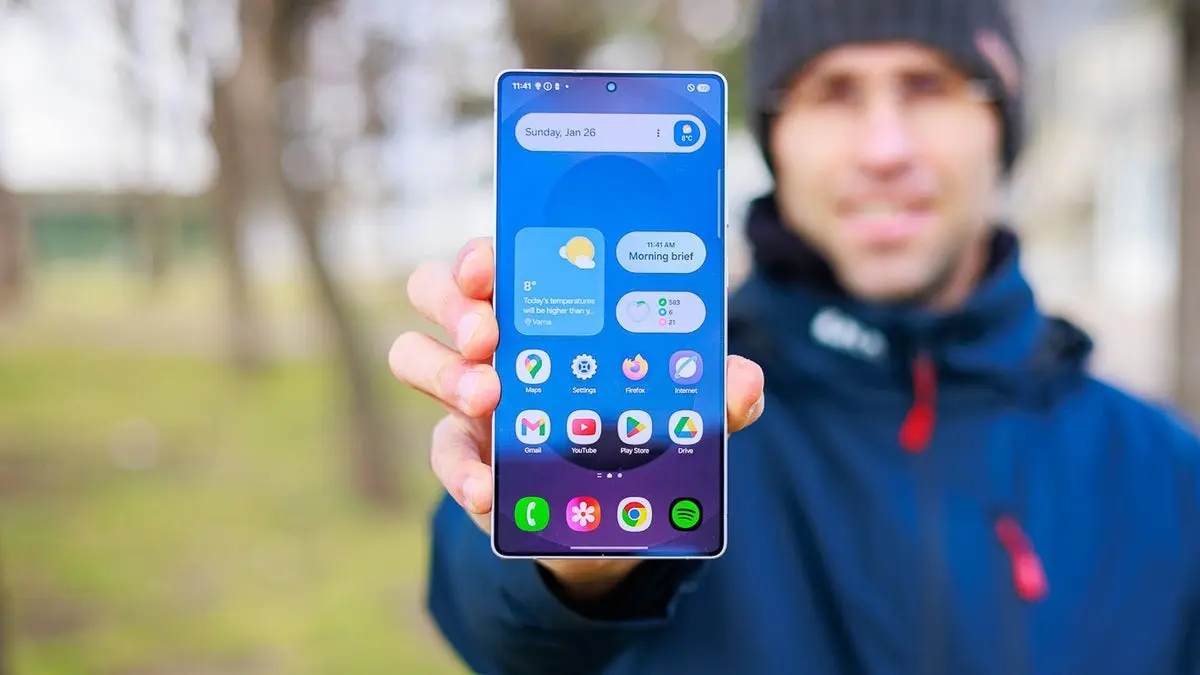 فردی در فضای باز که گوشی هوشمند را به سمت دوربین در دست گرفته و صفحهٔ اصلی را نشان می‌دهد.